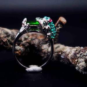 Könnte beinhalten: Ein silberner Ring mit einem grünen und blauen Juwelen-Frosch-Design. Der Ring liegt auf einem Stück Baumrinde.