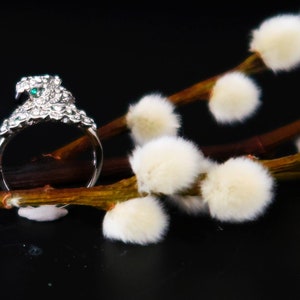 Könnte beinhalten: Ein silberner Ring mit einem grünen Stein und weißen Strasssteinen, vor einem Hintergrund aus flauschigen weißen Weidenzweigen.