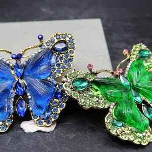 Könnte beinhalten: Zwei mit Juwelen besetzte Schmetterlingsbroschen. Eine ist blau mit passenden blauen und klaren Kristallen, die andere ist grün mit grünen und klaren Kristallen. Beide haben goldfarbene Metallarbeiten.