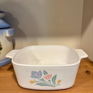 Puede incluir: Fuente de horno rectangular blanca con asas, decorada con un estampado floral en colores pastel. El diseño presenta flores azules, rosas y amarillas con hojas verdes. La fuente es de cerámica y está sobre una estantería de madera.