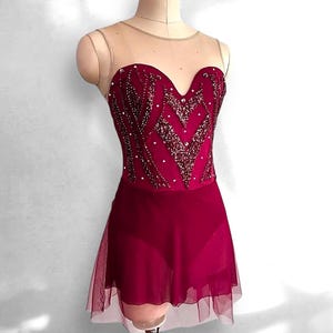 May include: A deep magenta leotard featuring a sweetheart neckline with a sheer nude mesh yoke. The bodice is embellished with intricate dark red sequin patterns and scattered clear crystals. A matching sheer magenta skirt overlays the base.