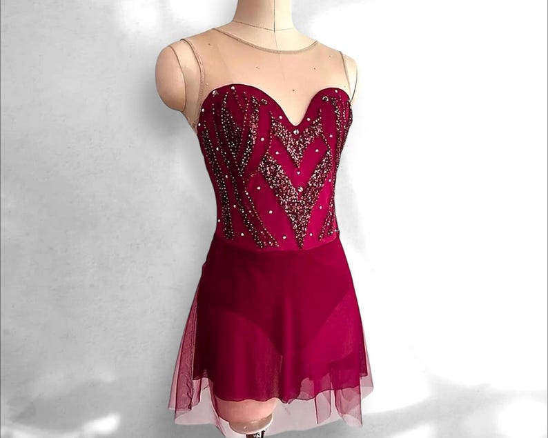 May include: A deep magenta leotard featuring a sweetheart neckline with a sheer nude mesh yoke. The bodice is embellished with intricate dark red sequin patterns and scattered clear crystals. A matching sheer magenta skirt overlays the base.