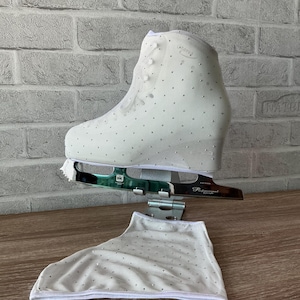 May include: A white ice skate boot adorned with numerous small crystals, featuring a metallic blade labeled MK Professional. A matching white fabric skate cover with crystal embellishments is positioned below the skate on a stand.