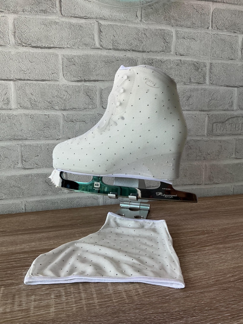 May include: A white ice skate boot adorned with numerous small crystals, featuring a metallic blade labeled MK Professional. A matching white fabric skate cover with crystal embellishments is positioned below the skate on a stand.