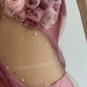 May include: A dance leotard featuring a nude mesh bodice adorned with scattered clear crystals and a cluster of layered fabric roses in shades of dusty pink and mauve. The garment includes sheer long sleeves and additional fabric roses at the waist.