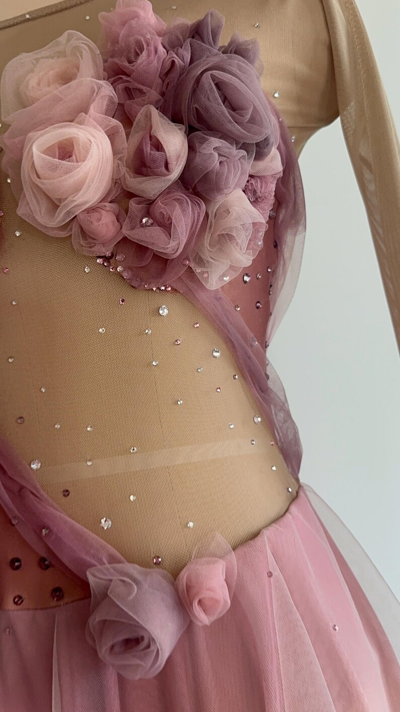 May include: A dance leotard featuring a nude mesh bodice adorned with scattered clear crystals and a cluster of layered fabric roses in shades of dusty pink and mauve. The garment includes sheer long sleeves and additional fabric roses at the waist.