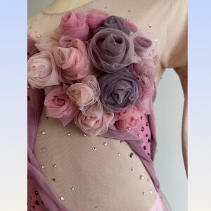 May include: A sleeveless garment featuring a nude-colored mesh bodice adorned with scattered clear crystals and a cluster of layered fabric roses in shades of dusty pink, mauve, and muted purple, with additional fabric draping along the side.
