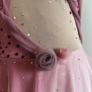 May include: A pink and mauve dance dress featuring a sheer nude mesh side panel adorned with scattered clear crystals. The garment is embellished with clusters of fabric roses in varying shades of pink and mauve, along with a layered tulle skirt.