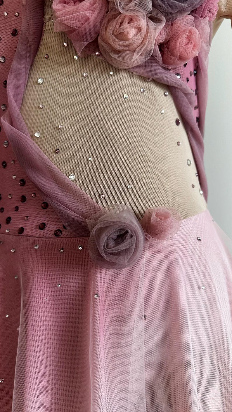 May include: A pink and mauve dance dress featuring a sheer nude mesh side panel adorned with scattered clear crystals. The garment is embellished with clusters of fabric roses in varying shades of pink and mauve, along with a layered tulle skirt.
