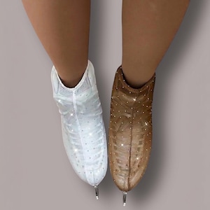 May include: A pair of ice skates featuring a sheer, mesh-like outer covering adorned with small, reflective crystals. One skate is white, and the other is a tan-brown color. Both skates have a pointed toe design and a metallic blade at the base.