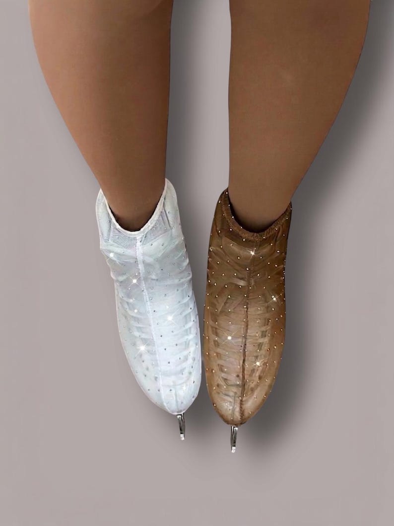 May include: A pair of ice skates featuring a sheer, mesh-like outer covering adorned with small, reflective crystals. One skate is white, and the other is a tan-brown color. Both skates have a pointed toe design and a metallic blade at the base.
