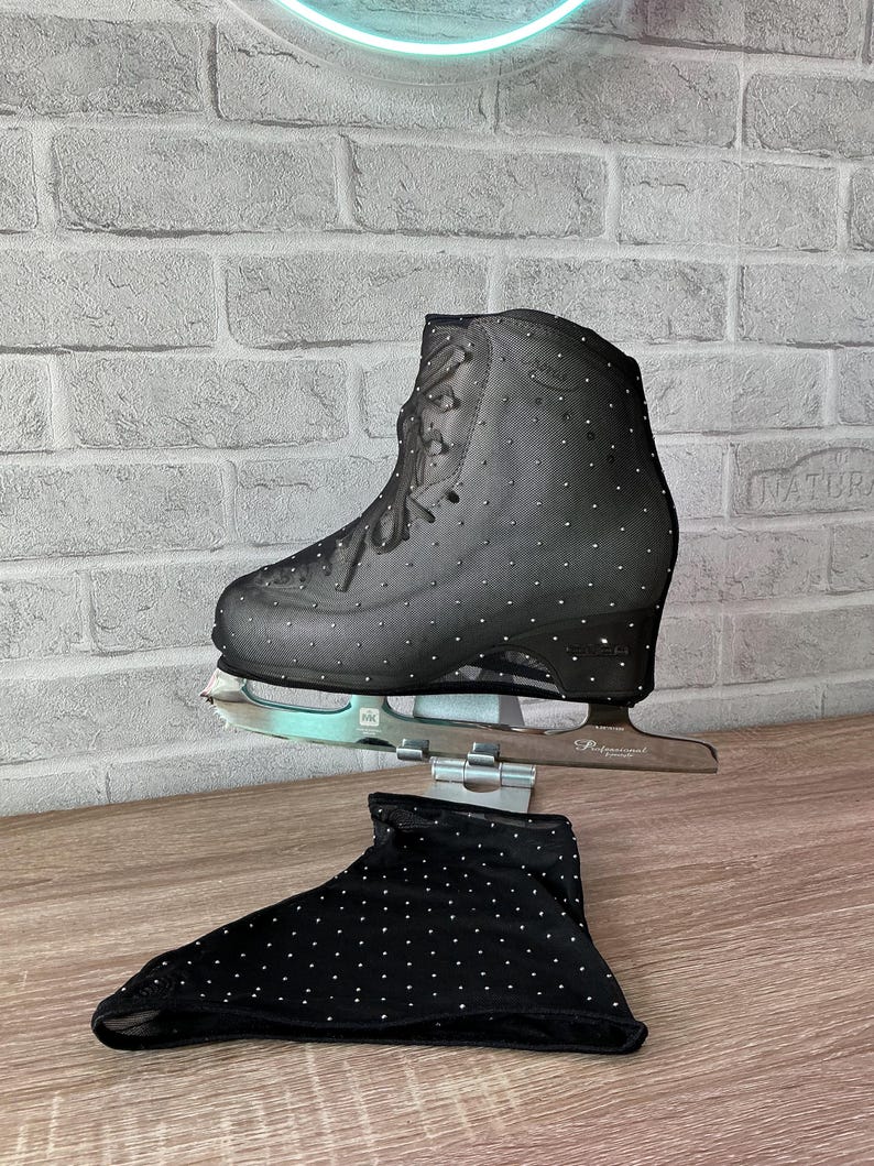 May include: A black ice skate featuring a mesh overlay adorned with small silver rhinestones, paired with a matching black fabric skate cover also decorated with silver rhinestones. The skate has a silver metal blade attached to the base.