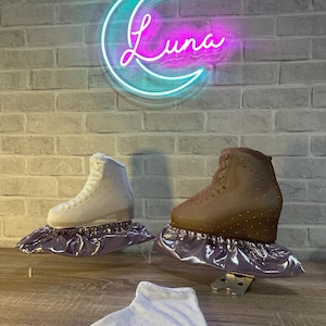 May include: Two ice skates, one white and one brown, each adorned with small crystals and resting on a metallic purple fabric cover. A separate white fabric cover with crystals lies in the foreground. A neon sign shaped like a crescent moon with the text Luna glows in blue and pink above.