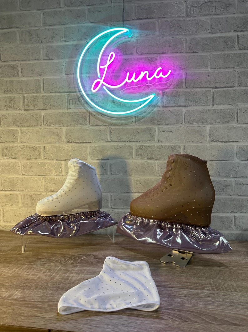 May include: Two ice skates, one white and one brown, each adorned with small crystals and resting on a metallic purple fabric cover. A separate white fabric cover with crystals lies in the foreground. A neon sign shaped like a crescent moon with the text Luna glows in blue and pink above.