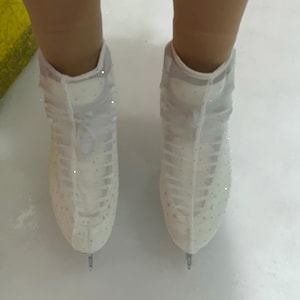 May include: A pair of white ice skates featuring sheer mesh covers adorned with small, sparkling crystals. The skates have a high-top design with white laces and are positioned on a smooth, white ice surface, showing the metal blades at the bottom.