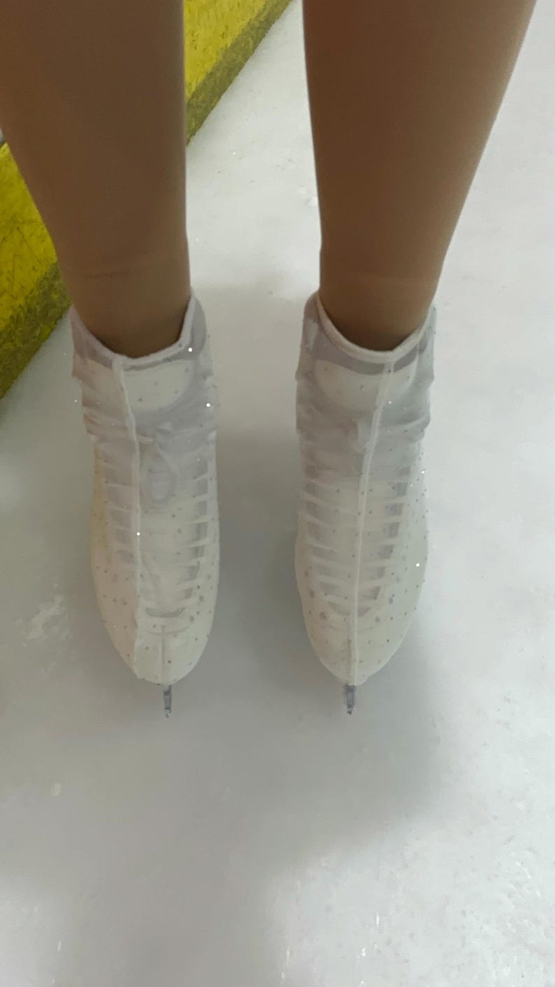 May include: A pair of white ice skates featuring sheer mesh covers adorned with small, sparkling crystals. The skates have a high-top design with white laces and are positioned on a smooth, white ice surface, showing the metal blades at the bottom.