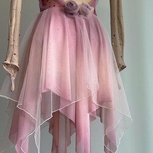 May include: A long-sleeved dance dress featuring a nude-colored bodice with scattered crystals and a layered, handkerchief-style skirt in shades of dusty pink. The waist is accented with two fabric roses, and the skirt has a white hemline.
