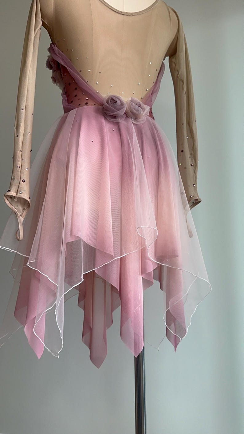 May include: A long-sleeved dance dress featuring a nude-colored bodice with scattered crystals and a layered, handkerchief-style skirt in shades of dusty pink. The waist is accented with two fabric roses, and the skirt has a white hemline.