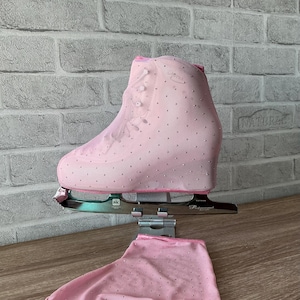 May include: A light pink ice skate cover featuring a mesh fabric adorned with small silver rhinestones. The cover is designed to fit over a figure skate boot, with a matching pink mesh fabric piece laid flat underneath the metal skate blade.