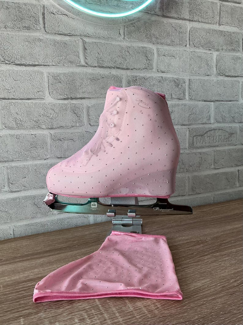 May include: A light pink ice skate cover featuring a mesh fabric adorned with small silver rhinestones. The cover is designed to fit over a figure skate boot, with a matching pink mesh fabric piece laid flat underneath the metal skate blade.