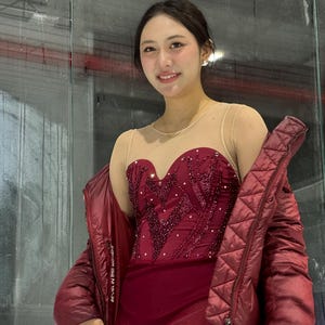 May include: A maroon figure skating dress with a sweetheart neckline, sheer nude mesh bodice, and intricate beadwork. The dress is paired with a matching maroon quilted puffer jacket featuring the text REVEL IN THIS MOMENT on the inner lining.