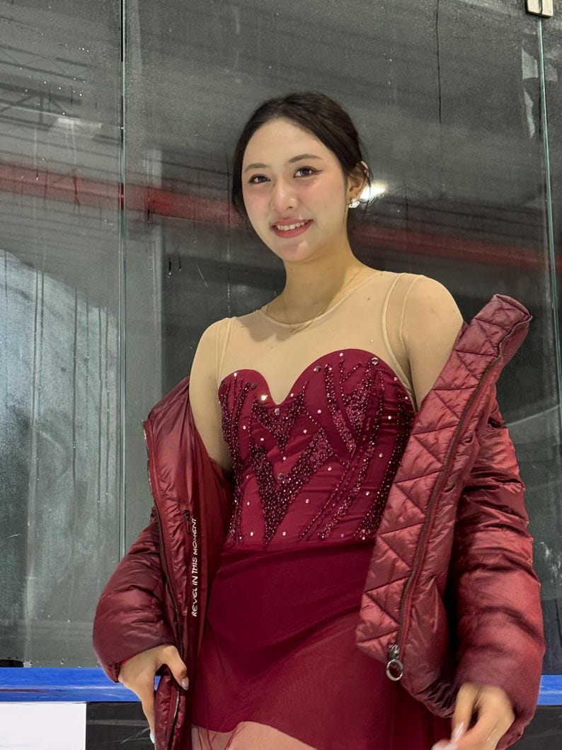 May include: A maroon figure skating dress with a sweetheart neckline, sheer nude mesh bodice, and intricate beadwork. The dress is paired with a matching maroon quilted puffer jacket featuring the text REVEL IN THIS MOMENT on the inner lining.