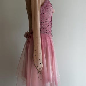 May include: A pink performance dress featuring a nude-colored long sleeve, a bodice embellished with scattered dark crystals, and a layered tulle skirt with a handkerchief hemline. The garment includes a small fabric flower detail at the waist.