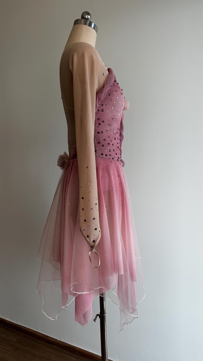 May include: A pink performance dress featuring a nude-colored long sleeve, a bodice embellished with scattered dark crystals, and a layered tulle skirt with a handkerchief hemline. The garment includes a small fabric flower detail at the waist.