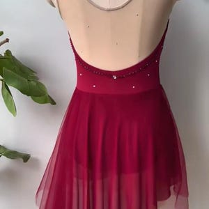 May include: A deep red dance leotard featuring a sheer beige mesh upper back and neckline, adorned with scattered small crystals. The garment includes a low-cut back design and an asymmetrical, layered tulle skirt that falls to a point on one side.