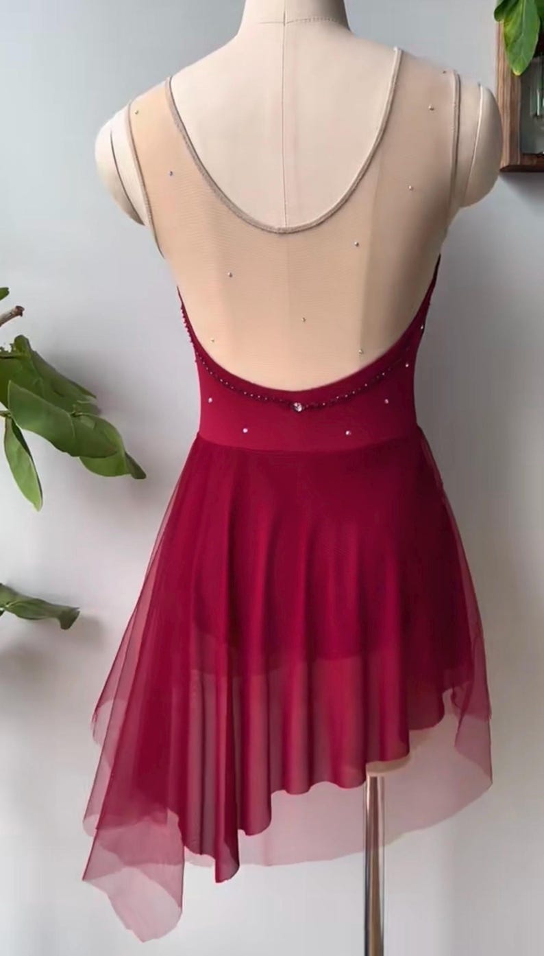 May include: A deep red dance leotard featuring a sheer beige mesh upper back and neckline, adorned with scattered small crystals. The garment includes a low-cut back design and an asymmetrical, layered tulle skirt that falls to a point on one side.