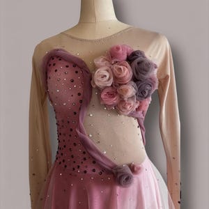 May include: A long-sleeved leotard featuring a nude mesh bodice with a pink ombre effect, adorned with scattered clear crystals. A cluster of fabric roses in shades of pink and mauve is attached to the upper chest, accented by draped fabric.