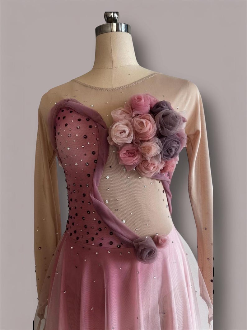 May include: A long-sleeved leotard featuring a nude mesh bodice with a pink ombre effect, adorned with scattered clear crystals. A cluster of fabric roses in shades of pink and mauve is attached to the upper chest, accented by draped fabric.