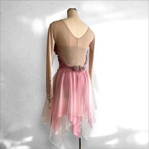 May include: A long-sleeved dance dress featuring a nude-colored mesh bodice with a V-neckline, adorned with scattered rhinestones. The skirt consists of layered pink tulle with an asymmetrical hem, accented by two small fabric roses at the waist.