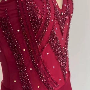May include: A deep red sleeveless bodice featuring a sweetheart neckline, embellished with vertical rows of dark red sequins and scattered clear crystals. The garment is constructed from a smooth fabric with a sheer overlay at the top section.