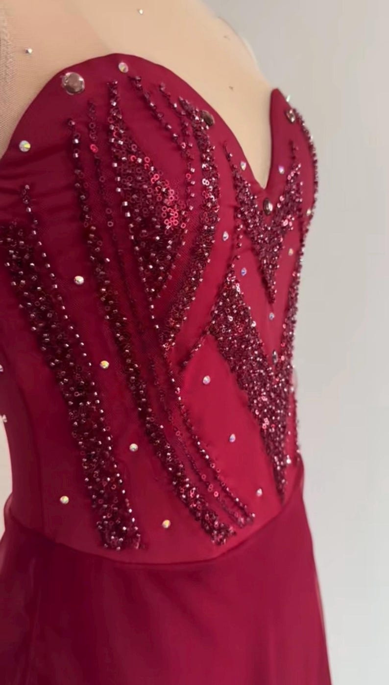 May include: A deep red sleeveless bodice featuring a sweetheart neckline, embellished with vertical rows of dark red sequins and scattered clear crystals. The garment is constructed from a smooth fabric with a sheer overlay at the top section.