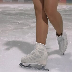 May include: A pair of white figure skates with metallic blades, featuring a lace-up design and subtle sparkling embellishments on the boot exterior, worn on an ice rink surface.