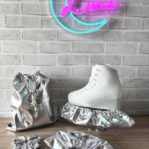 May include: A white ice skate with small embedded crystals, accompanied by a metallic silver drawstring bag, two elasticised silver skate covers, and a matching silver scrunchie. The items are displayed against a brick wall with a neon moon sign.