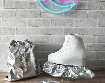 May include: A white ice skate with small embedded crystals, accompanied by a metallic silver drawstring bag, two elasticized silver skate covers, and a matching silver scrunchie. The items are displayed against a brick wall with a neon moon sign.