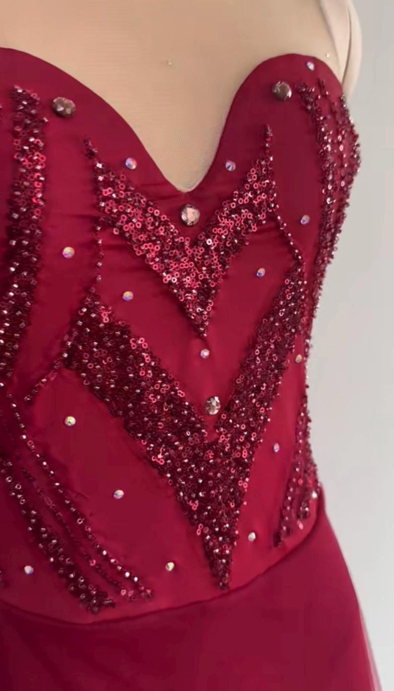 May include: A deep red bodice featuring a sweetheart neckline with a sheer mesh insert. The fabric is heavily embellished with dark red sequins arranged in chevron patterns, accented by scattered clear crystals throughout the front surface.