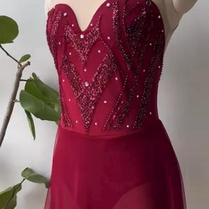 May include: A deep red leotard featuring a sweetheart neckline and a sheer nude mesh upper bodice. The bodice is embellished with intricate dark red sequin patterns and scattered crystals. A sheer red skirt overlay is attached at the waist.