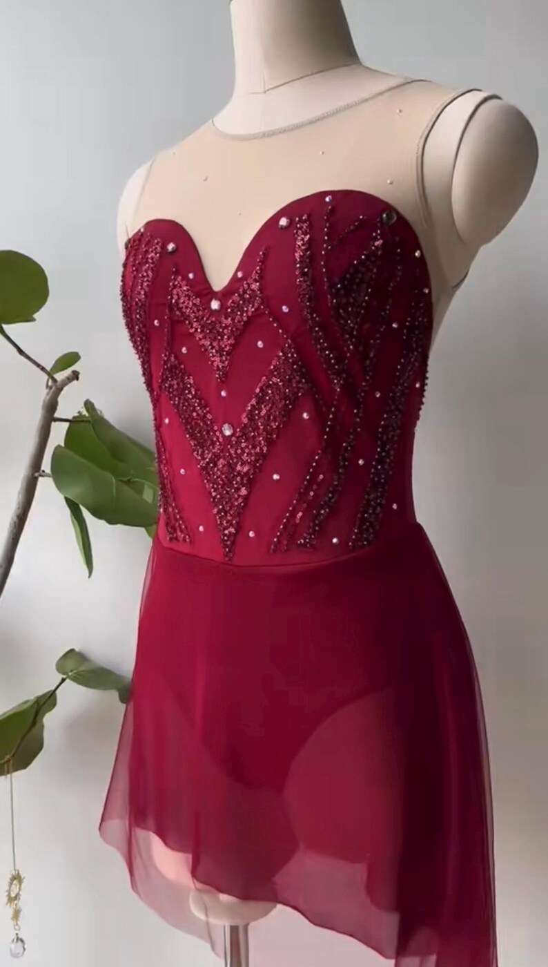May include: A deep red leotard featuring a sweetheart neckline and a sheer nude mesh upper bodice. The bodice is embellished with intricate dark red sequin patterns and scattered crystals. A sheer red skirt overlay is attached at the waist.