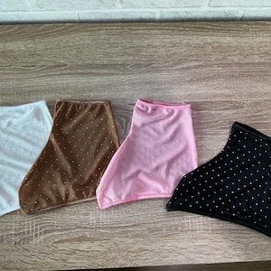 May include: Four fabric neck gaiters in white, brown, pink, and black. Each gaiter features a repeating pattern of small, evenly spaced white dots across the surface. The items are arranged side-by-side, showcasing their distinct colors.