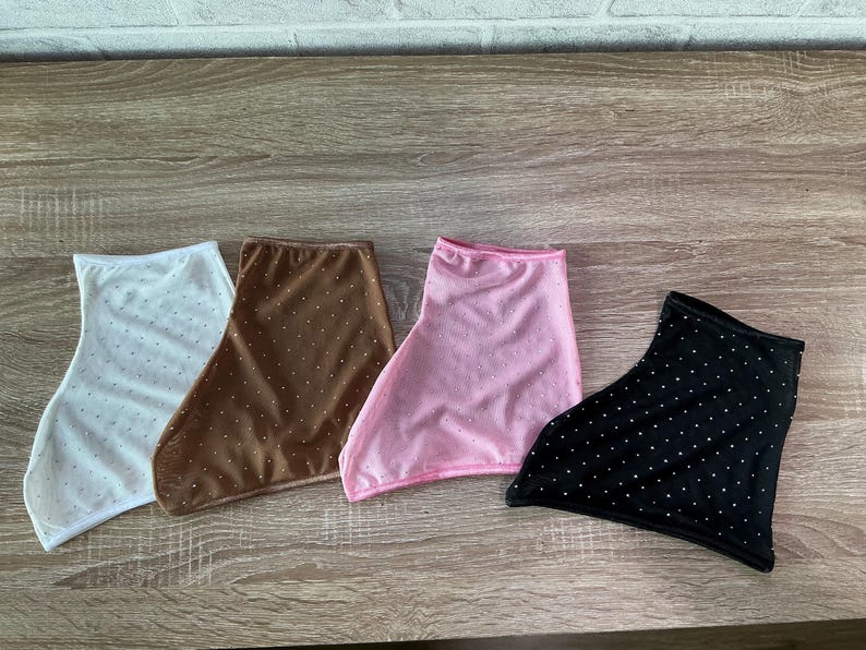 May include: Four fabric neck gaiters in white, brown, pink, and black. Each gaiter features a repeating pattern of small, evenly spaced white dots across the surface. The items are arranged side-by-side, showcasing their distinct colors.