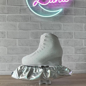 May include: A white ice skate with small embedded crystals on the boot, resting on a metallic silver elasticized blade cover. The skate is positioned in front of a neon sign shaped like a crescent moon with the word Luna written in script.