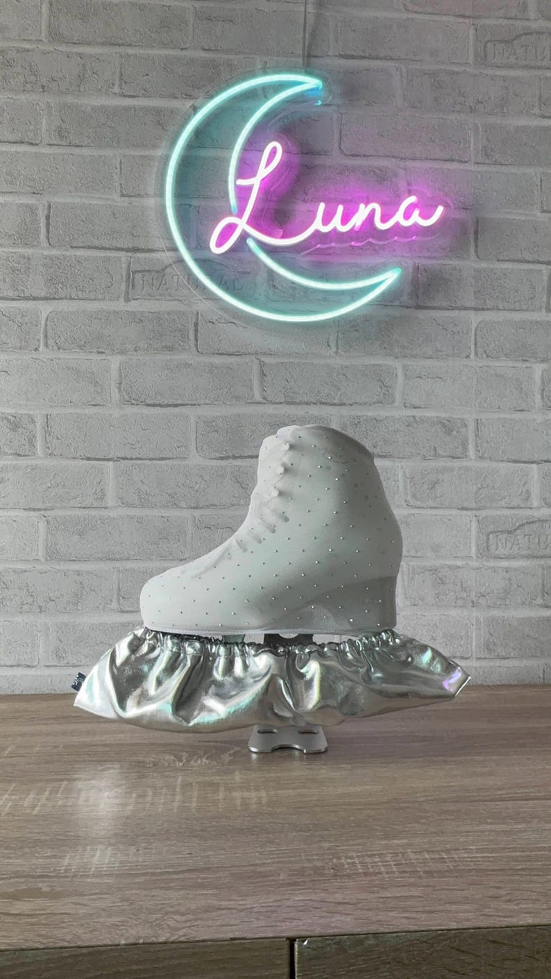 May include: A white ice skate with small embedded crystals on the boot, resting on a metallic silver elasticized blade cover. The skate is positioned in front of a neon sign shaped like a crescent moon with the word Luna written in script.