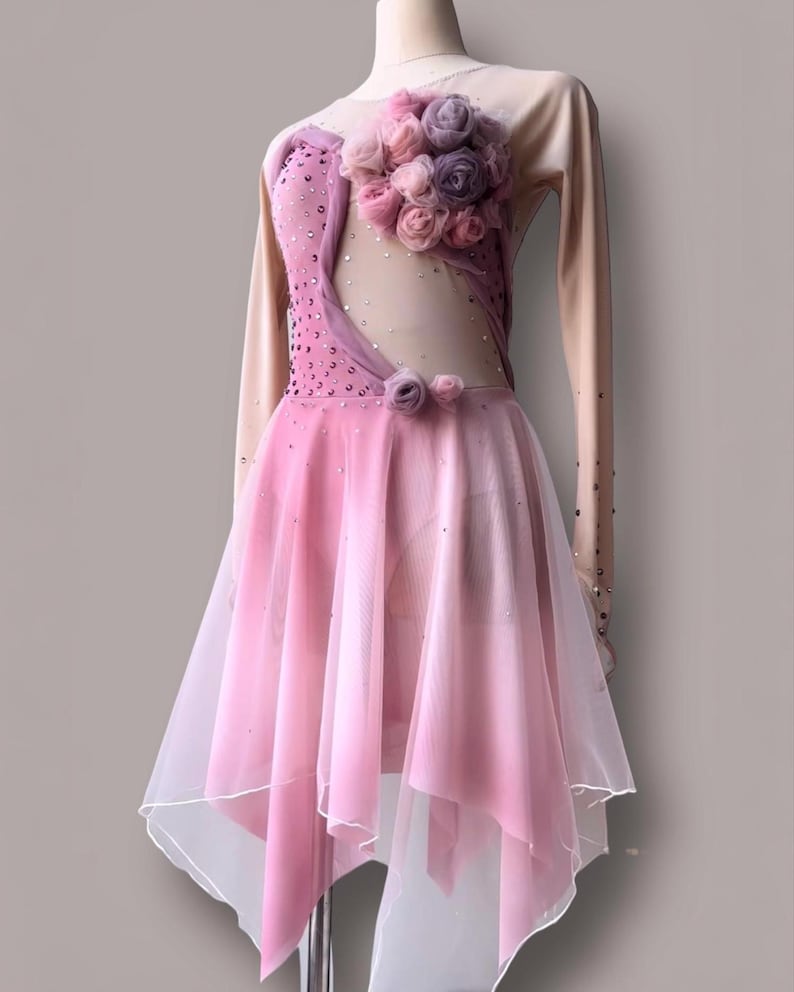 May include: A long-sleeved performance dress featuring a nude-colored mesh bodice with pink fabric panels, scattered crystals, and a cluster of pink and purple fabric roses on the chest. The skirt is made of layered pink sheer fabric.