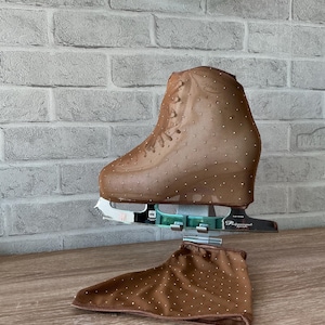 May include: A tan ice skate cover featuring a sheer mesh fabric adorned with small, evenly spaced silver rhinestones. The cover is fitted over a professional ice skate blade and boot, with a matching fabric piece resting below the skate.