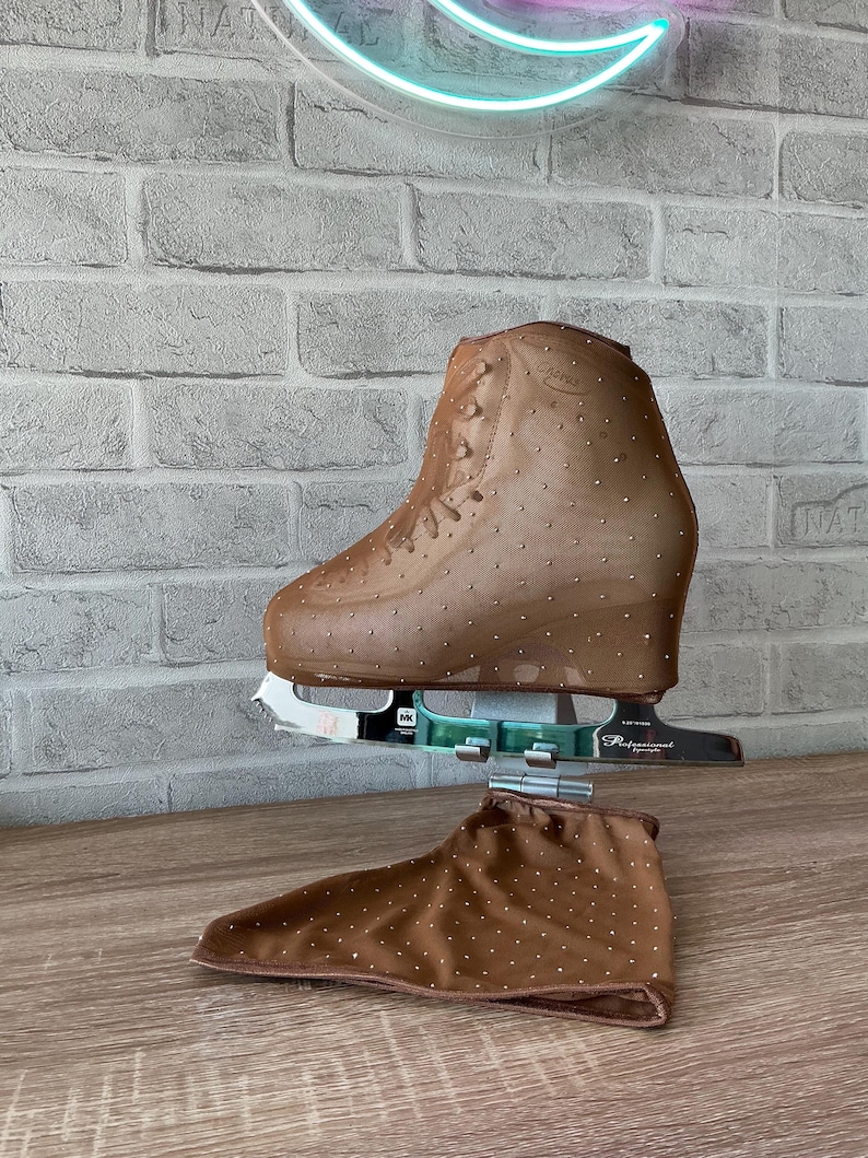 May include: A tan ice skate cover featuring a sheer mesh fabric adorned with small, evenly spaced silver rhinestones. The cover is fitted over a professional ice skate blade and boot, with a matching fabric piece resting below the skate.