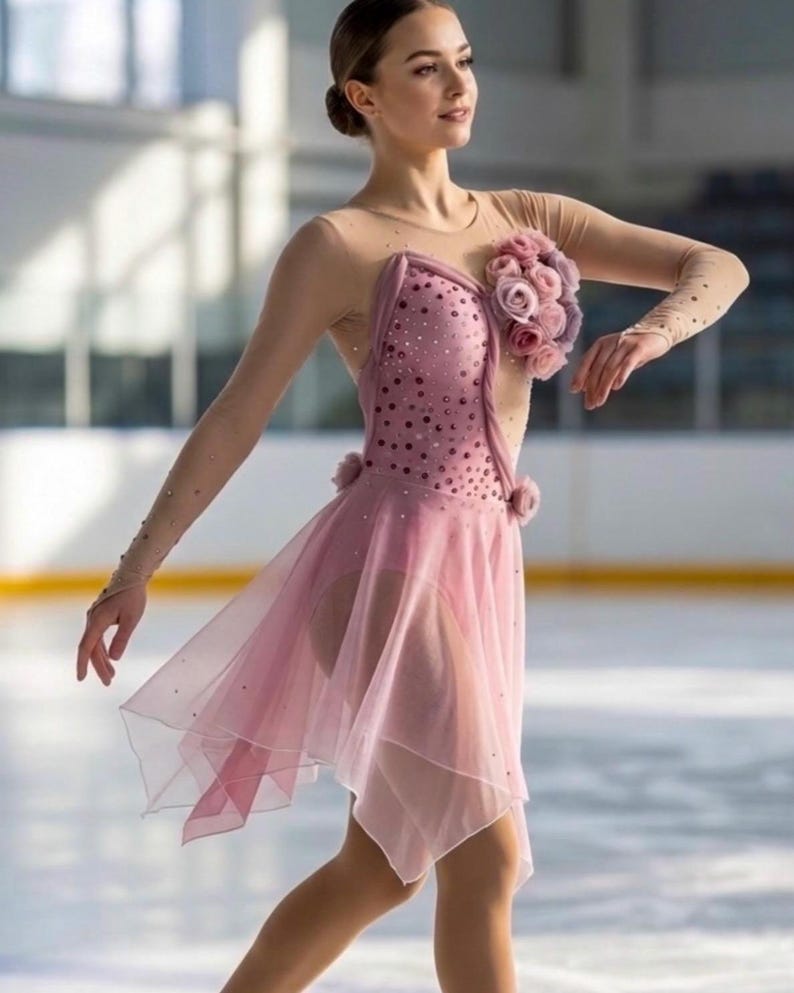 May include: A dusty pink figure skating dress featuring a sheer nude mesh bodice with long sleeves, a bodice adorned with scattered dark pink crystals, and a cluster of fabric roses on the left shoulder. The skirt is made of layered sheer fabric.