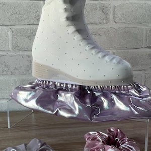 May include: A white ice skate boot covered in small silver crystals, resting on a metallic lavender skate cover. Two matching metallic lavender scrunchies are positioned in front of the skate on the surface.